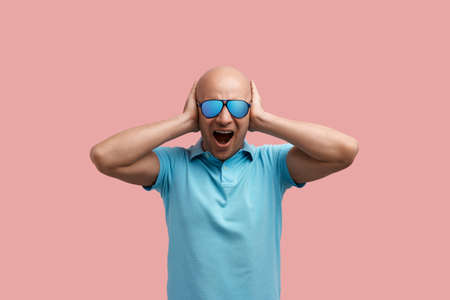 Displeased bald unshaven homosexual man is screaming, annoyed with loud music and noise, closes ears with hands.の写真素材