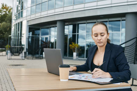 European business woman is working with laptop outdoors during coffee break, plans presentation, is focused on documents, looks through printed charts, wears business suit and deep neckline blouseの写真素材
