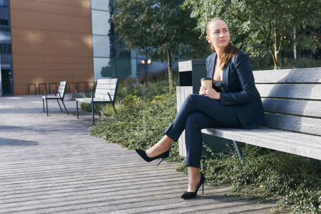 Pensive european business woman has coffee break, sits outside of office on bench with takeaway cup in hand, looks aside, wears business suit, high heels and deep neckline blouse, has ponytailの写真素材