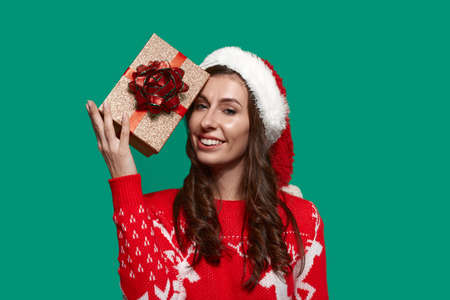 Smiling brunette is touching forehead with golden gift box with bow knot, trying to guess what is...