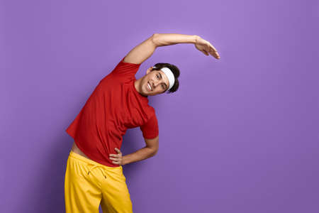 Smiling handsome sportsman is stretching, bends over with raised arm, keeps hand on waist, coaching...