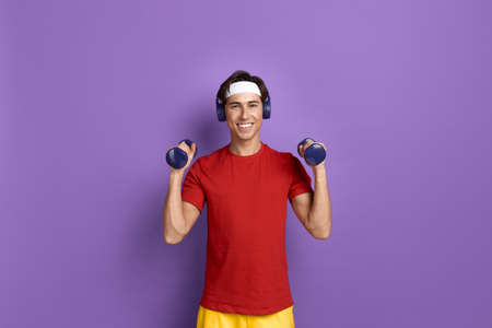 Smiling handsome fitness instructor is raising hands with dumbbells, happy to work out in gym, wears...