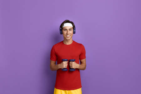 Smiling handsome fitness instructor holds dumbbells in front of chest, happy to work out in gym,...