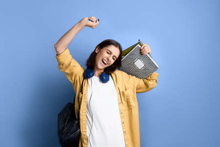 Happy student girl passed exam, is stretching in relief with closed eyes, smiles broadly, holding books, has bright manicure, wearing yellow shirt, white t-shirt, black bag and headphones over neckの写真素材
