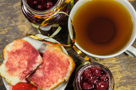Tea with cherry jam. Red jam on white bread, tea, red spoons on old wooden backgroundの写真素材