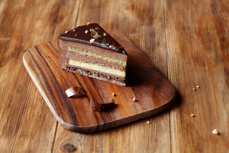 Piece of layered Chocolate Hazelnut Mousse Cake covered with chocolate glaze and decorated with chocolate elements, on wooden board, on wooden background.の写真素材