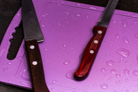 Two knives lie on a cutting board in a dark sink in the kitchen. The board is covered with drops of...