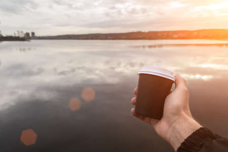 A cardboard cup of coffee in a mans hand on the background of a pond. Coffee to go.の写真素材