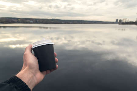 A cardboard cup of coffee in a mans hand on the background of a pond. Coffee to go.の写真素材