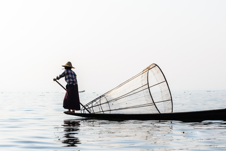 Burmese fisherman on bamboo boat catching fish in traditional way with handmade net. Inle lake, Myanmar (Burma) travel destinationのeditorial素材