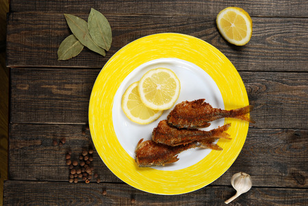fried fish with lemon, round pepper and bay leaf on a plate on a wooden backgroundの写真素材