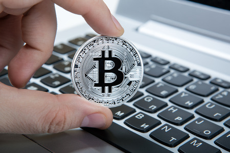 hand holds a silver coin of bitcoin above the keyboardの写真素材