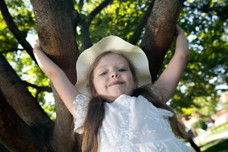 little girl in a white dress and panama sits on a tree portraitの写真素材