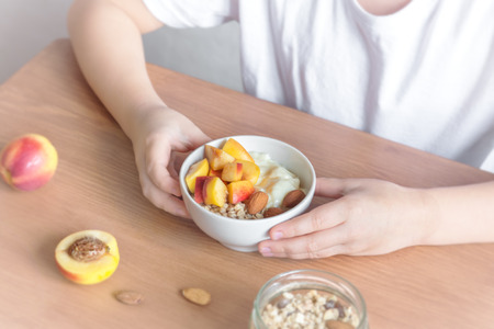 Eating healthy breakfast bowl. Oatmeal granola, yougurt, fresh nectarins inwhite bowl in female (child) hands over wooden table. Clean eating, dieting, detox, vegetarian, vegan food concept.の写真素材