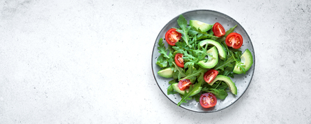 Green salad with sliced avocado, tomatoes and fresh arugula on white stone table with copy space, banner. Healthy diet vegan vegetarian summer vegetable salad food concept.の写真素材