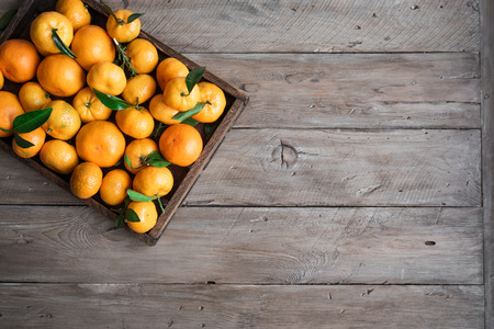 Tangerines (oranges, clementines, citrus fruits) with green leaves in box on wooden background, copy space.の写真素材