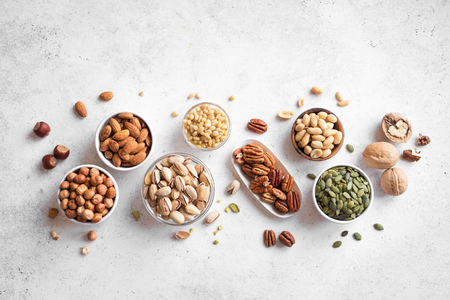 Various Nuts in  bowls on white background, top view, copy space. Nuts assortment - pecans, hazelnuts, walnuts, pistachios, almonds, pine nuts, peanuts, pumpkin seeds.の写真素材