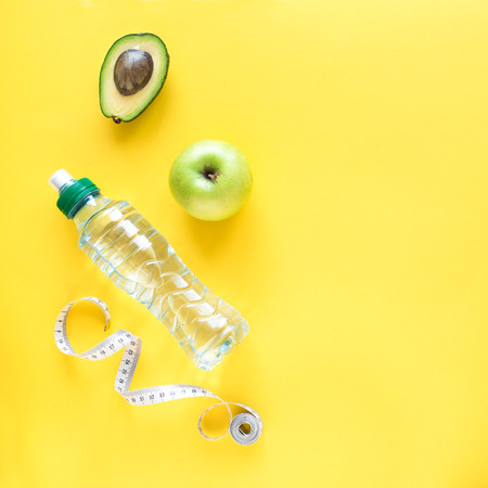 Healthy diet concept on yellow, top view, copy space. Measuring tape, green apple, avocado and water...