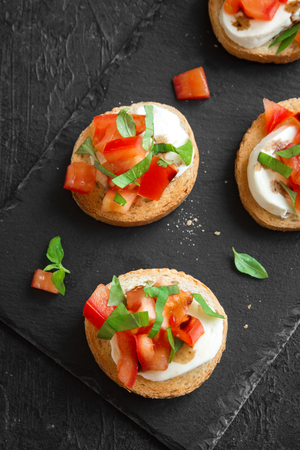 Italian bruschetta with chopped tomatoes, basil, mozzarella cheese and balsamic vinegar. Fresh homemade caprese bruschetta or crostini over black stone background.の写真素材
