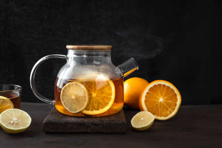 Citrus tea with lemon, oranges in transparent glass teapot on dark wooden table, copy space. Fruit tea. Seasonal winter autumn hot drink.の写真素材