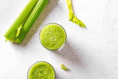 Celery juice in glasses. Fresh squeezed detox green juice and bunch of organic celery on white table, top view, copy space.の写真素材