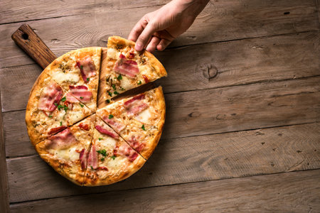 Hand picking slice of bacon pizza. Fresh homemade italian bacon and cheese pizza on wooden...