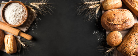 Assortment of various delicious freshly baked bread and flour ingredient on black background, banner, copy space. Variety of artisan bread and ears of wheat.の写真素材