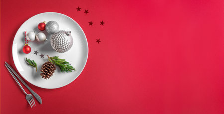 Beautiful Christmas Table Setting. Festive silver baubles and pine cone on white plate on red background, creative flat lay, banner, copy space. Christmas dinner, party design, menu concept.の写真素材