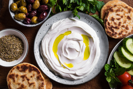 Labneh yogurt cream cheese with olive oil, zaatar, olives, vegetables and pita bread on wooden table, close up. Traditional arabian or middle eastern breakfast with homemade labneh dip.の写真素材