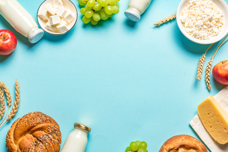 Happy Shavuot - Religious Jewish holiday concept with dairy products, cheese, traditional bread, milk bottles, wheat and grapes on blue background, frame, copy space.の写真素材