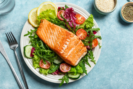 Grilled salmon fish fillet and fresh vegetable salad with tomato, lettuce, arugula. Healthy food - green salad and roasted salmon on blue background.の写真素材