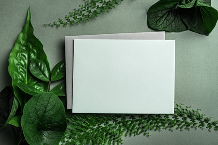 Greeting or invitation blank card and green plant leaves on dark green background. Stylish creative mock up, top view, copy spaceの写真素材