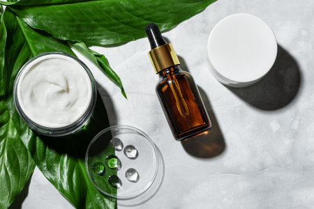 Natural cosmetics for skin care and green plant leaves on marble background, flat lay. Bottle of serum, essential oil, face cream jar. Eco beauty concept.の写真素材