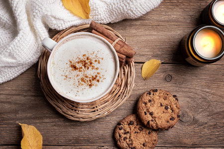 Autumn cozy composition. Cup of cappuccino coffee, chocolate chip cookies, burning candles and yellow autumn leaves on wooden background, top view. Hygge comfortable concept flat lay.の写真素材