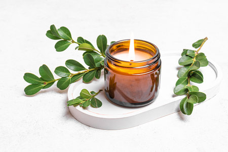 Burning soy wax candle in amber glass jar and green plant leaves on white concrete tray. Natural home decor, spa template and aroma massage concept.の写真素材