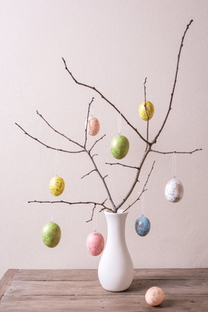 Painted Easter eggs on twigs in white vase, neutral background, minimal composition. Happy Easter...