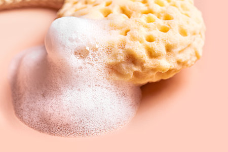 Face sponge and white soap foam on pink background close up. Body care and hygiene concept.