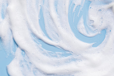 White soft foam texture swatch on blue pastel background. Face cleanser mousse sample, soap foam.
