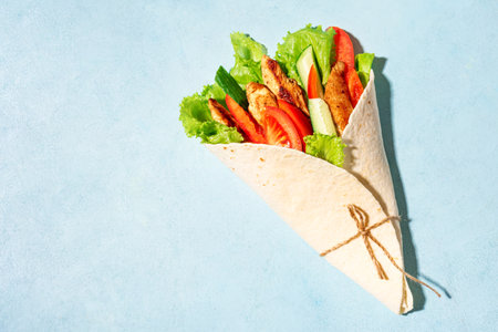 Grilled chicken and vegetables salad tortilla wrap on blue background, top view, copy space. Fresh...