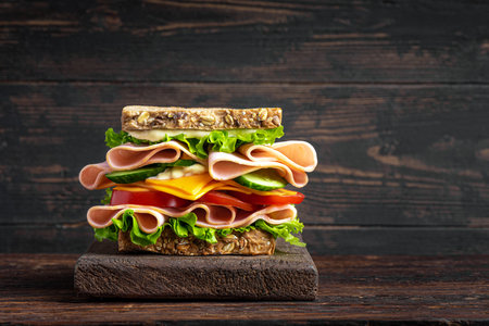Big layered sandwich with ham, lettuce, cheddar cheese, tomato and cucumber on dark wooden background, copy space. Healthy homemade lunch: sandwich with wholegrain bread, meat, vegetables and cheese.の写真素材