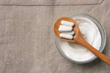 Probiotic capsules on wooden spoon and bowl of kefir or yogurt on linen napkin, top view, copy...