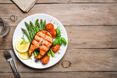 Grilled salmon steak with green asparagus, tomatoes and lemon slices on wooden background, top view, copy space. Balanced healthy meal, keto lunch.の写真素材