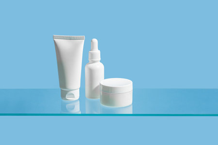 Cosmetic products containers on glass shelf and blue background, copy space. White blank face moisturizer jar, cleanser gel tube, face serum bottle branding, daily skin care routine.の写真素材