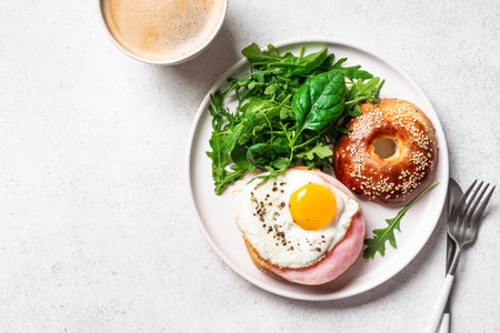Bagel sandwich with fried egg, cream cheese, ham and greens and cup of coffee. Healthy homemade balanced breakfast, white background, top view, copy space.の写真素材