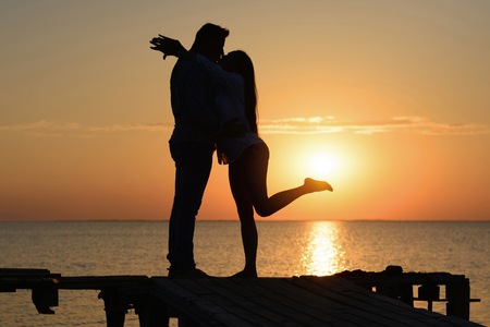 Silhouette of a kissing guy and a girl on a bridge by the ocean at sunset. Honey month (love and romance)の写真素材