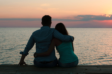 Two lovers (pair) in an embrace sit on the seashore and look at the sunset. Romance and Travelの写真素材