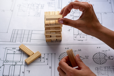 Man architect draws a plan, graph, design, geometric shapes by pencil on large sheet of paper at office desk and builds model house from wooden blocks (bars).の写真素材