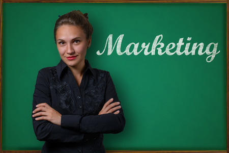 Beautiful young woman teacher (student, business woman) in classical dress standing near a blackboard with the inscription Marketingの写真素材