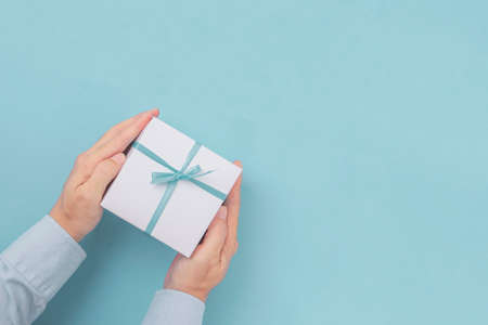Hands holding Gift box with blue bow with copy space in craft paper over blue background. Top view,...