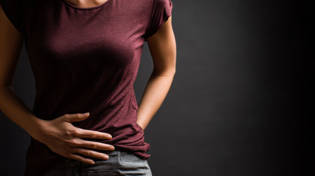 Woman holding stomach, dark background, maroon shirt, pain expression, health concept, digestive discomfort, emotional tensionの素材